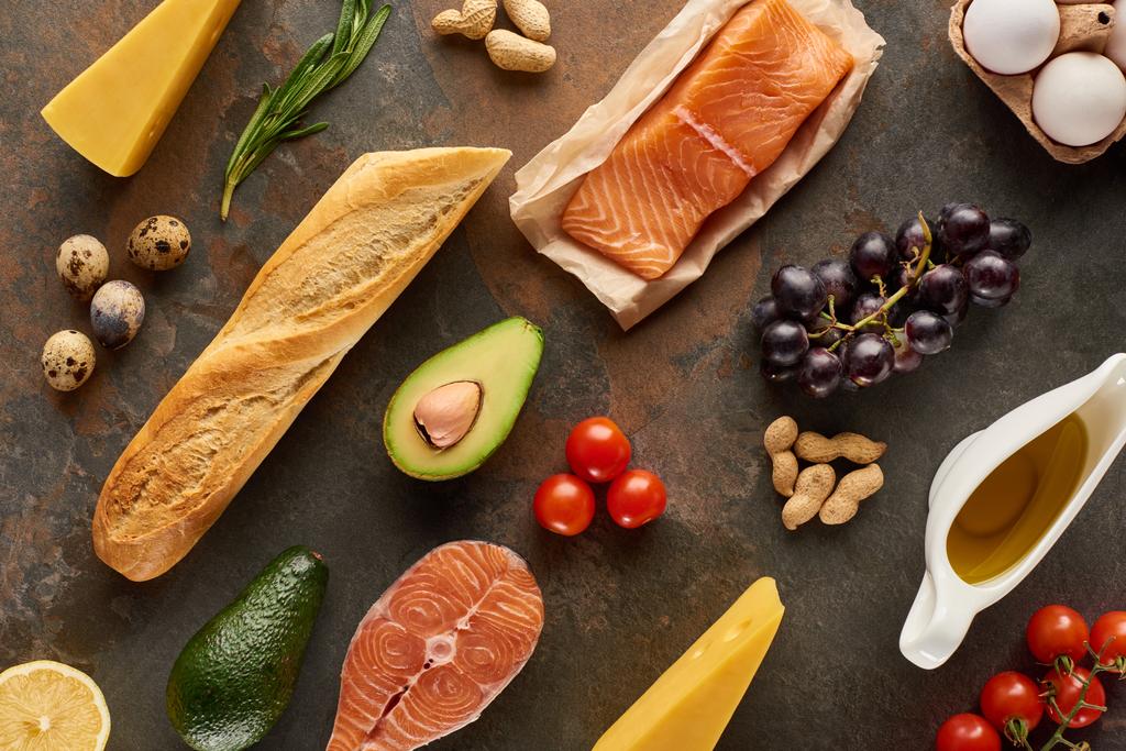Top uitzicht van rauwe vis in de buurt van groenten, fruit, eieren, kaas, pinda's, stokbrood en olijfolie op marmer oppervlak - Foto, afbeelding