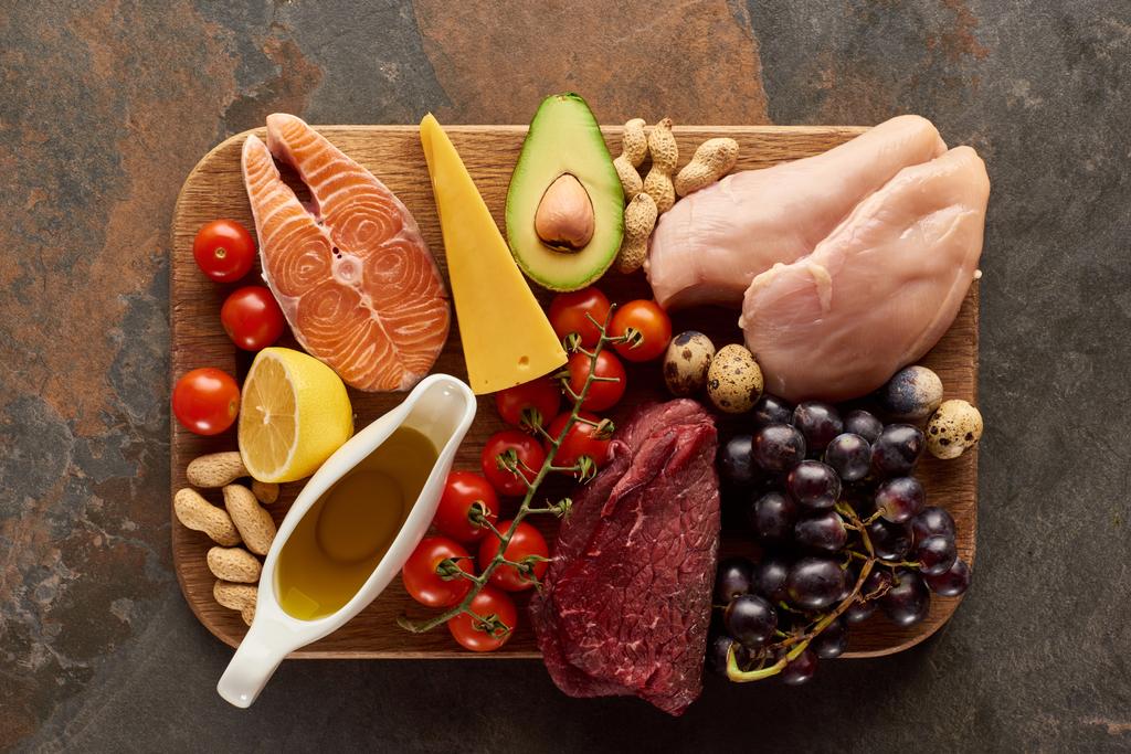 Top uitzicht van rauw vlees, vis, gevogelte, kaas, fruit, groenten met olijfolie en pinda's op houten snijplank op donker bruin marmeren oppervlak - Foto, afbeelding