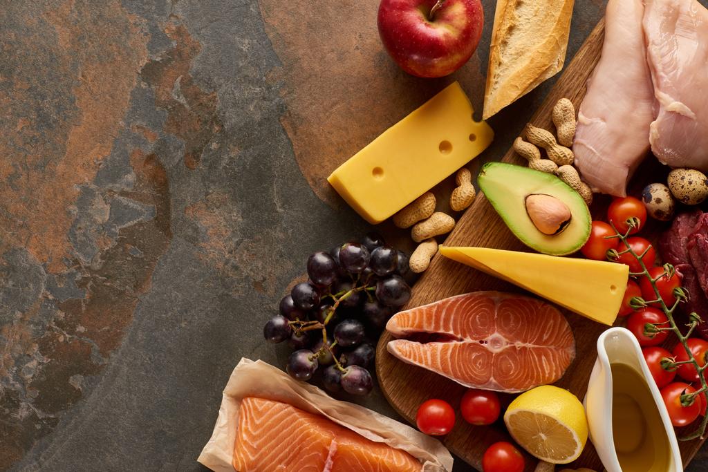Ansicht des Holzschneidebretts mit rohem Fisch, Geflügel, Käse, Obst, Gemüse, Baguette und Erdnüssen auf dunkelbrauner Marmoroberfläche mit Kopierraum - Foto, Bild