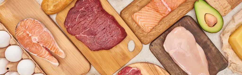 Panoramic shot raw meat, fish and poultry on wooden cutting boards near chicken eggs and half of avocado - Photo, Image
