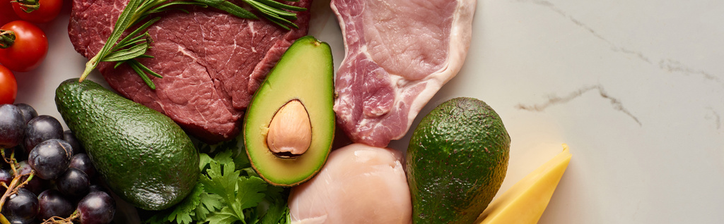 Panoramische foto van avocado's, rauw vlees, kipfilet, tomaten, druiven en kaas met groen op marmer oppervlak - Foto, afbeelding