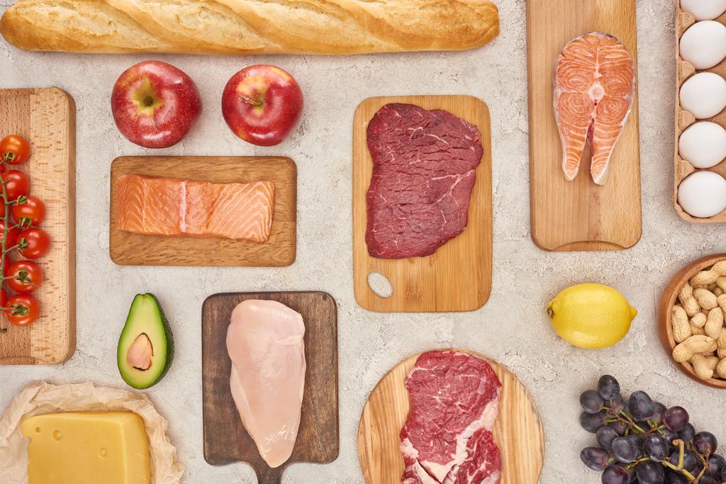 Top uitzicht van diverse vlees, gevogelte, vis, eieren, fruit, groenten, kaas en stokbrood op houten snijplanken op marmeren oppervlak - Foto, afbeelding