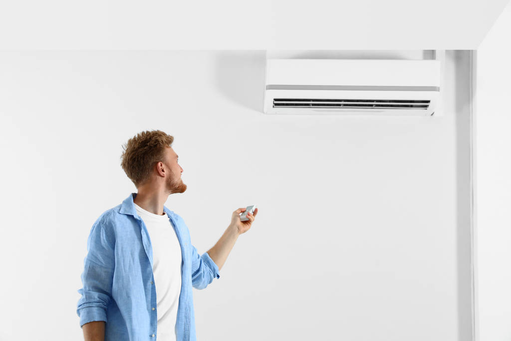 Junger Mann bedient Klimaanlage mit Fernbedienung in Innenräumen. Raum für Text - Foto, Bild
