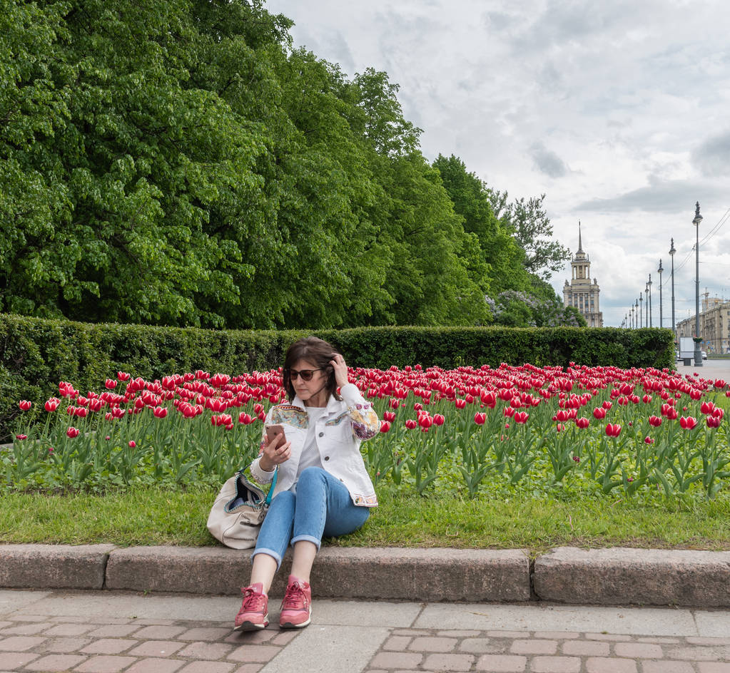 European woman sitting on a bed of tulips in the city Park and looking at the phone - Photo, Image