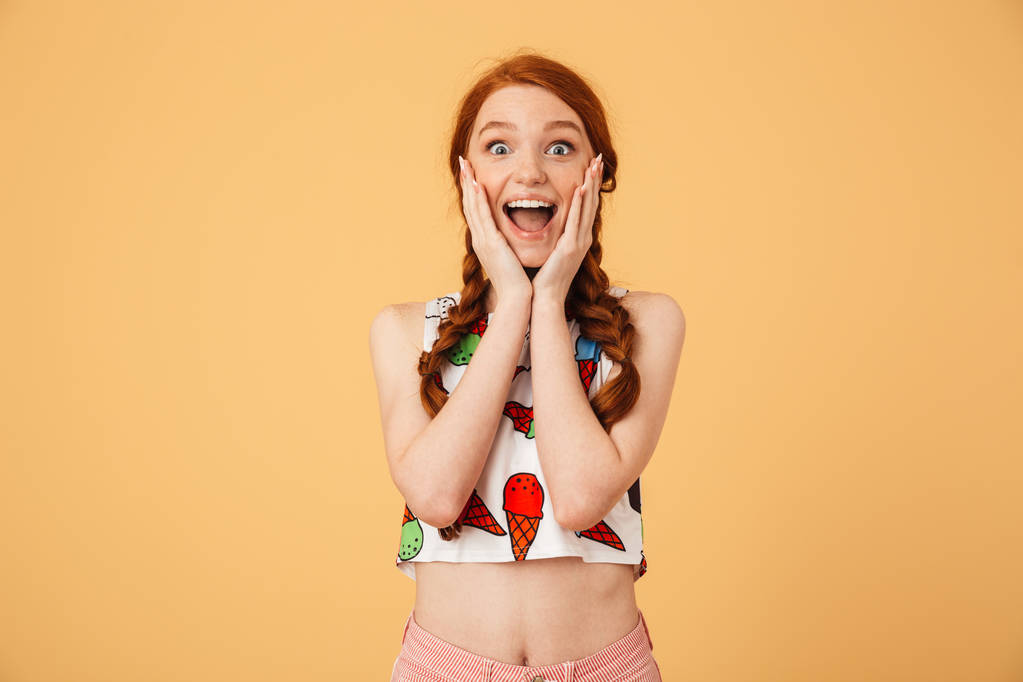 Ενθουσιασμένη όμορφη κοκκινομάλλα γυναίκα ντυμένη σε παγωτό τυπωμένο μπλουζάκι που παρουσιάζουν απομονωμένοι πάνω από κίτρινο φόντο. - Φωτογραφία, εικόνα