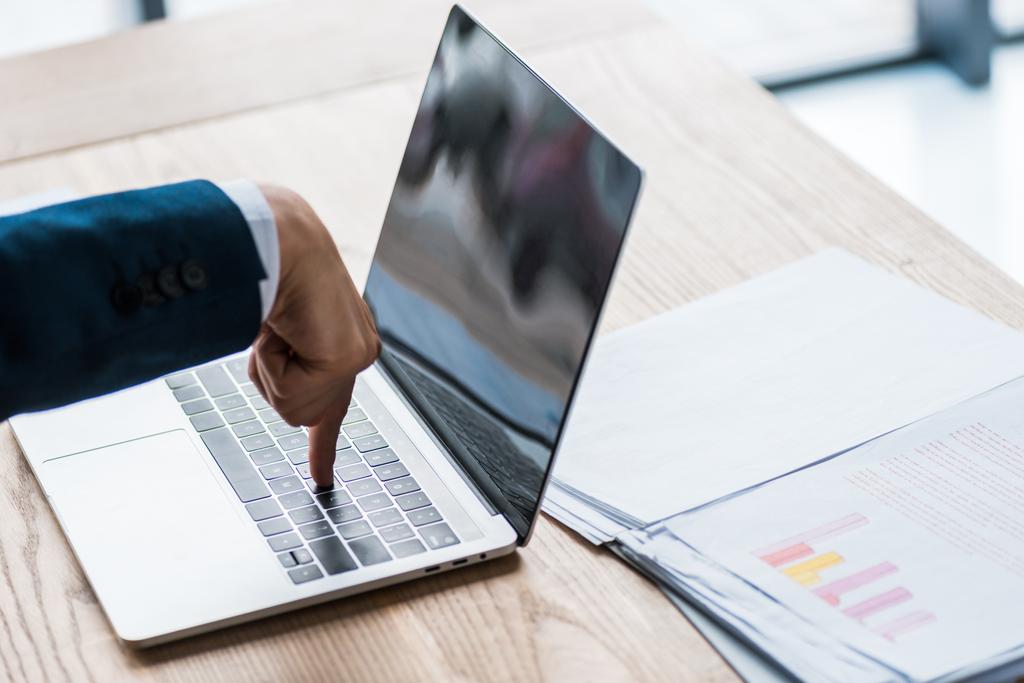 Ausgeschnittene Ansicht eines Mannes, der mit dem Finger auf die Laptop-Tastatur in der Nähe von Diagrammen und Diagrammen zeigt  - Foto, Bild