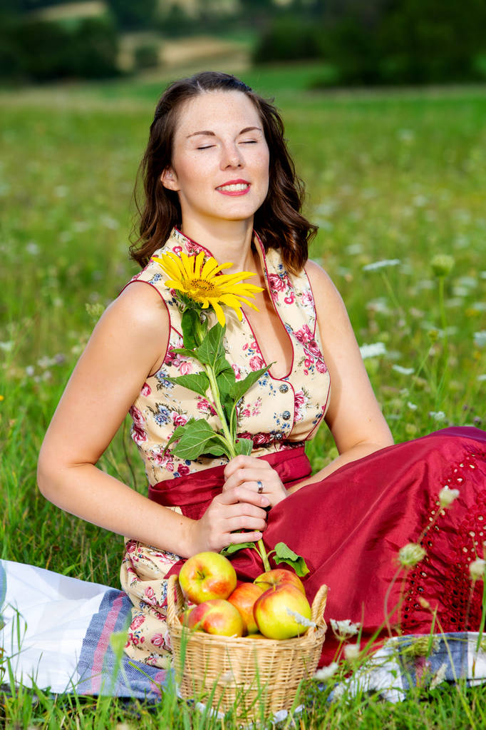 молода жінка в дирндлі сидить на лузі і тримає соняшник
 - Фото, зображення