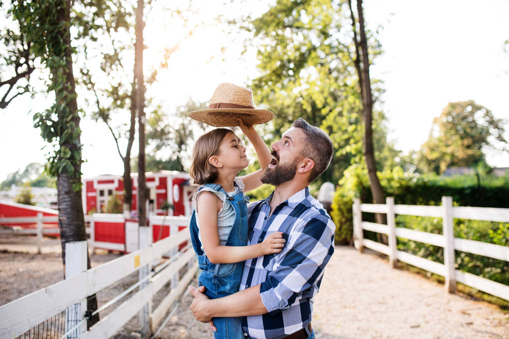 Un père avec une petite fille debout à l'extérieur sur la ferme familiale, jouant avec le chapeau
. - Photo, image
