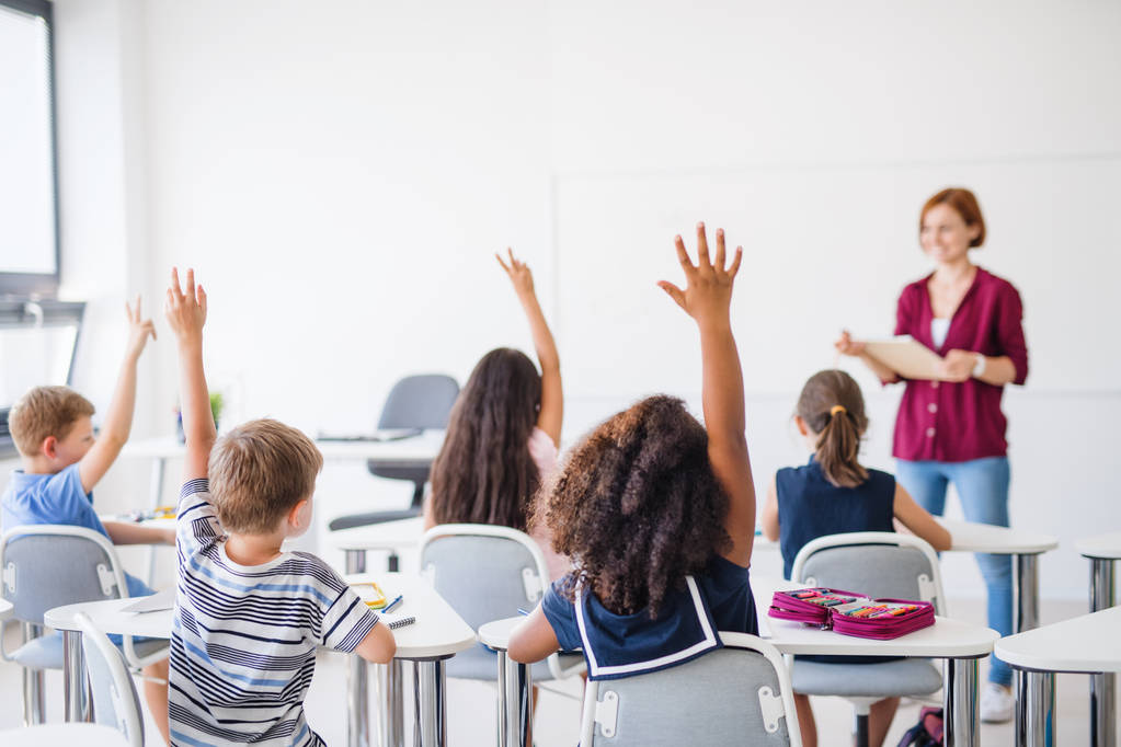 Vista trasera de los niños de la escuela sentados en el escritorio en el aula, levantando las manos
. - Foto, Imagen