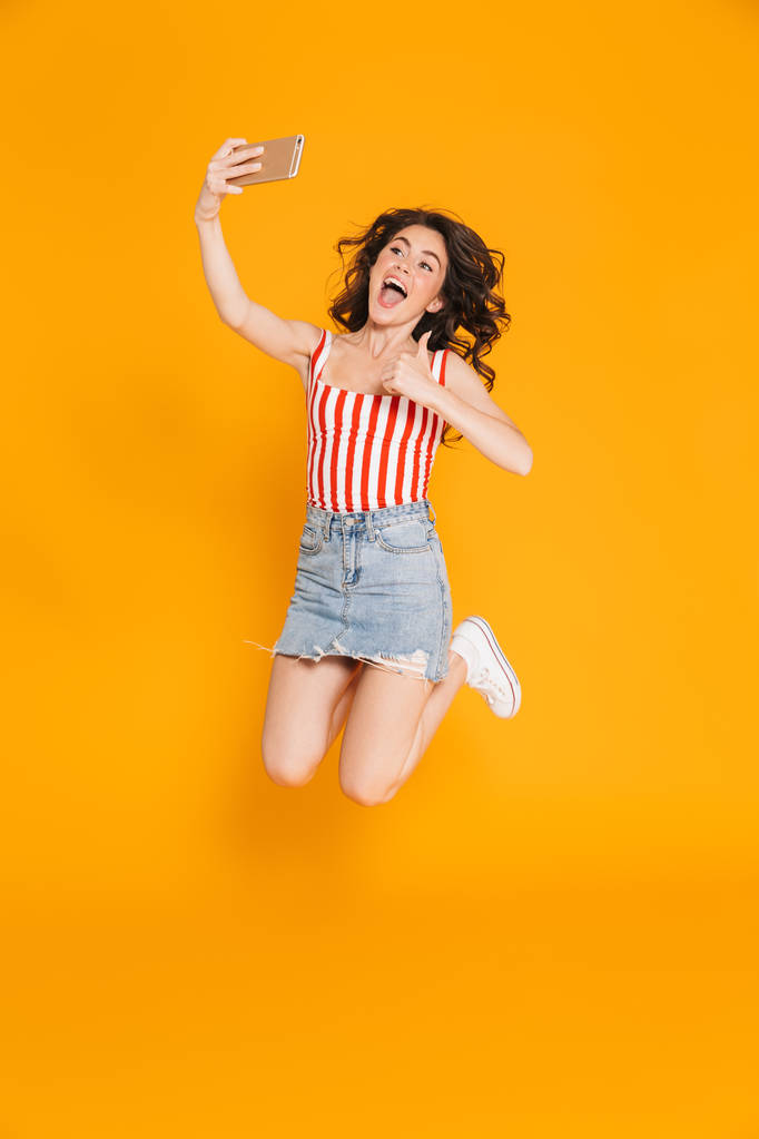 Portret podekscytowany brunetka Kobieta w denim spódnica skoki i pokazując kciuka w górę podczas robienia selfie na telefon komórkowy - Zdjęcie, obraz