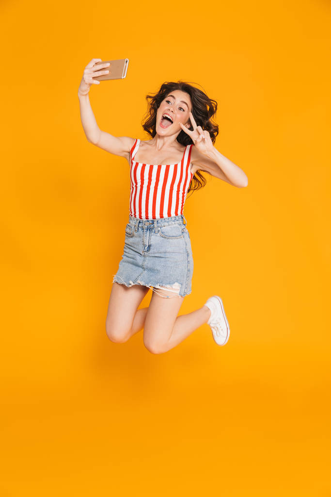 Muotokuva hämmästynyt brunette nainen denim hame hyppääminen ja osoittaa rauhaa laulaa ottaessaan selfie matkapuhelimeen
 - Valokuva, kuva