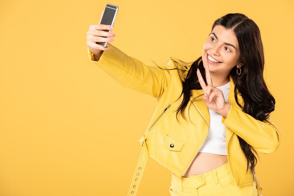 勝利のサインを示し、スマートフォンで自分撮りを取る美しい女の子は、黄色で隔離 - 写真・画像