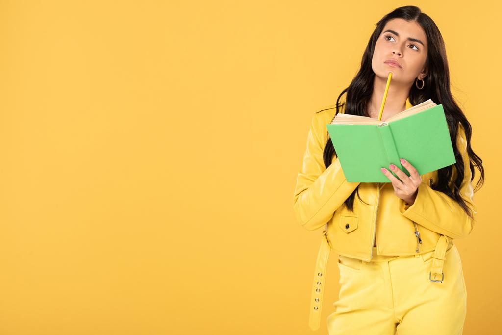 σκεπτική κορίτσι κρατώντας μολύβι και βιβλίο, απομονωμένο σε κίτρινο  - Φωτογραφία, εικόνα