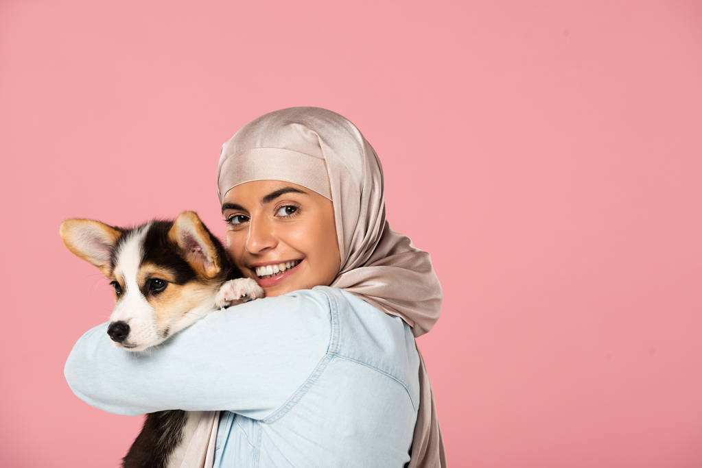 χαμογελαστή μουσουλμανική γυναίκα σε χιτζάμπ που κρατούν Corgi κουτάβι, απομονώνονται σε ροζ - Φωτογραφία, εικόνα
