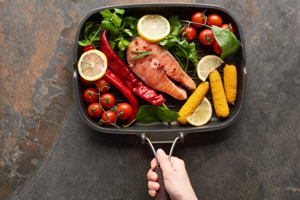 cropped view of woman holding raw salmon with vegetables, lemon and herbs in grill pan - Photo, Image