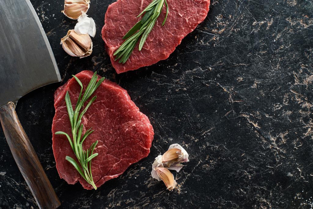 top view of raw beef steaks with rosemary twigs and garlic cloves near sharp knife on black marble surface - Photo, Image