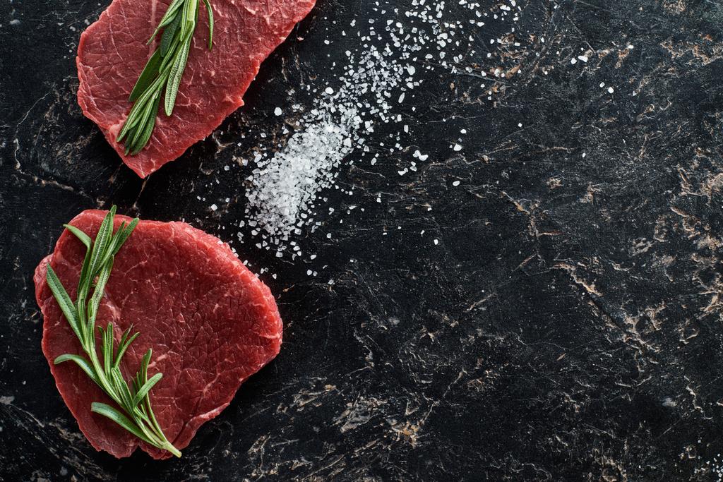 top view of raw beef steaks with rosemary twigs on black marble surface with scattered salt crystals - Photo, Image