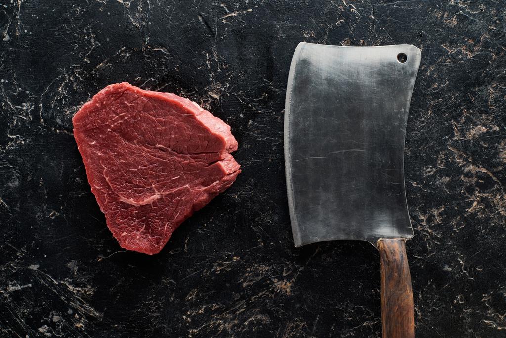 top view of butcher knife near raw beef steak on black marble surface - Photo, Image