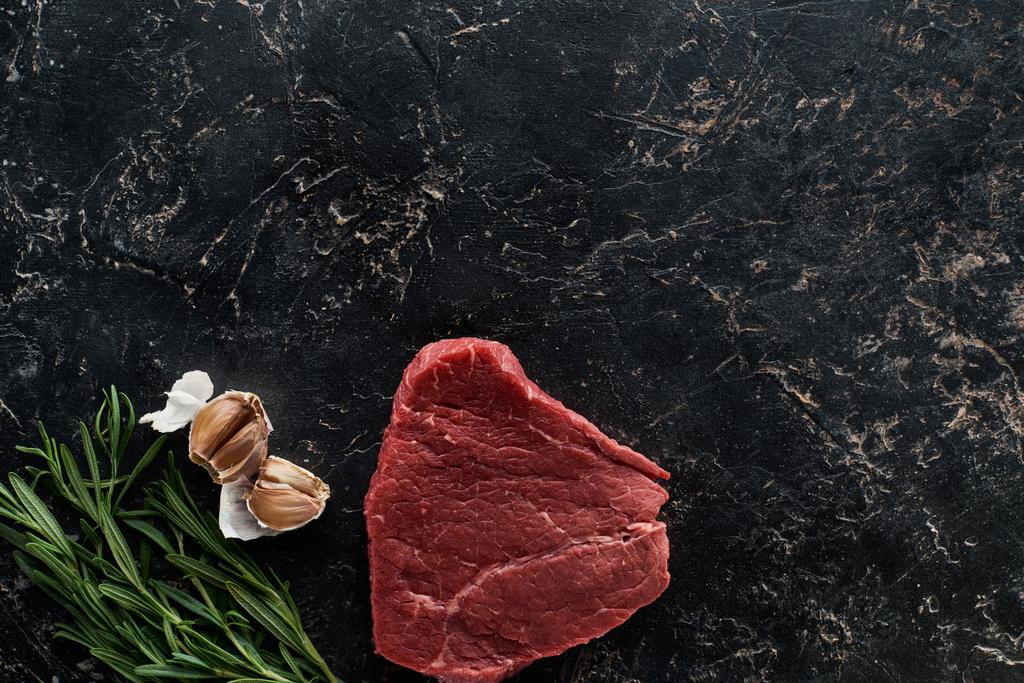 Top uitzicht van rauwe biefstuk in de buurt van rozemarijn twijgen en knoflook kruidnagel op zwart marmeren oppervlak - Foto, afbeelding
