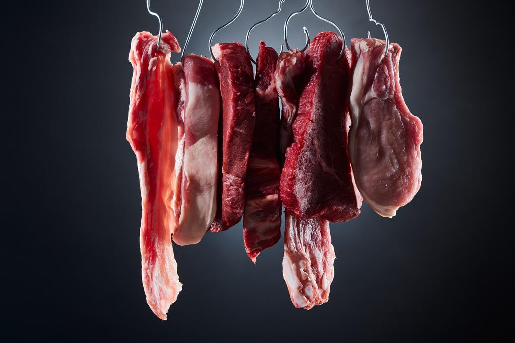 valikoituja raakoja lihapihvejä ja pekonia metallikoukuilla tummanmustalla pohjalla
 - Valokuva, kuva