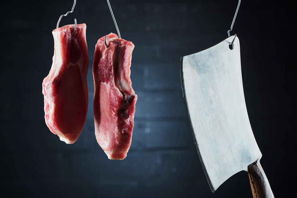 сирі свинячі стейки та м'ясний ніж на металевих гачках на темно-чорному тлі
 - Фото, зображення