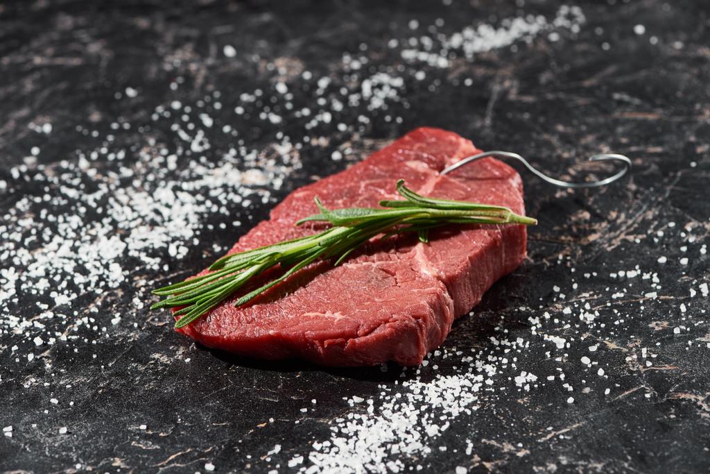 raw meat steak with rosemary twig on black marble surface with scattered salt  - Photo, Image