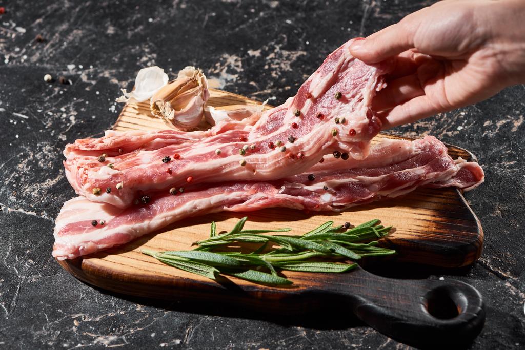 bijgesneden uitzicht van rauwe varkensvlees slice in de buurt van rozemarijn en knoflook op zwart marmeren oppervlak - Foto, afbeelding