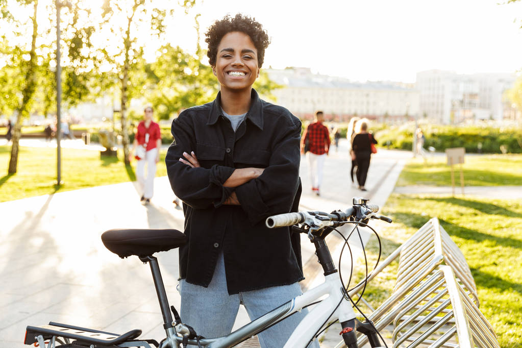 Photo of young african american woman standing with bicycle while walking in city park - Photo, Image