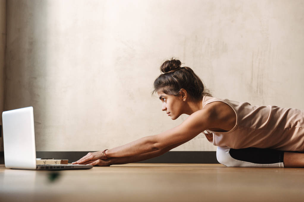 Foto de una mujer morena concentrada haciendo ejercicios de yoga usando un portátil mientras está sentada en el suelo en casa
 - Foto, Imagen