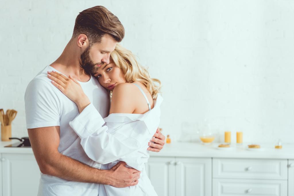 beautiful young woman looking at camera while embracing with boyfriend in kitchen - Photo, Image