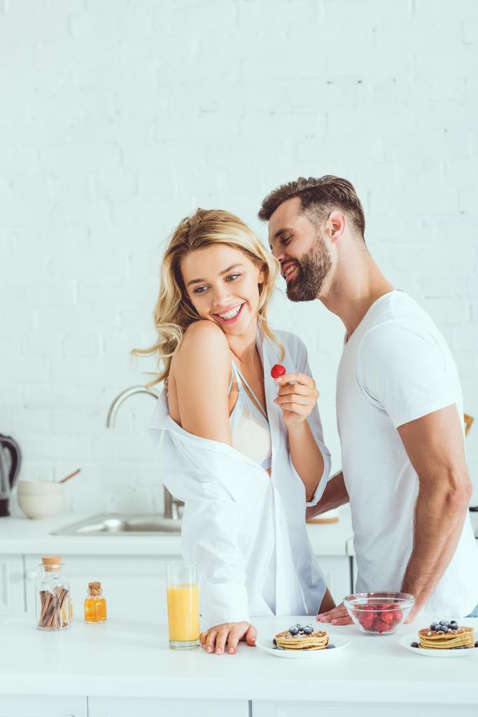 usměvavý muž objímající šťastnou přítelkyni s jahodovou v kuchyni - Fotografie, Obrázek