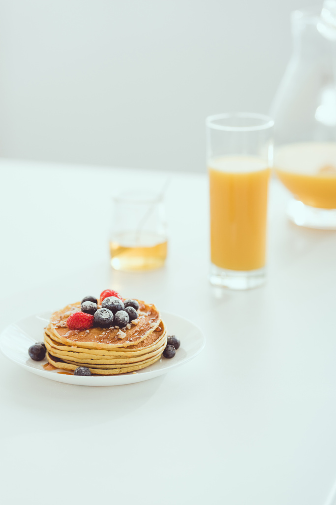 επιλεκτική εστίαση της πλάκας με νόστιμες τηγανίτες και μούρα κοντά σε γυαλί και κανάτα χυμό πορτοκαλιού και βάζο με μέλι σε λευκό τραπέζι - Φωτογραφία, εικόνα