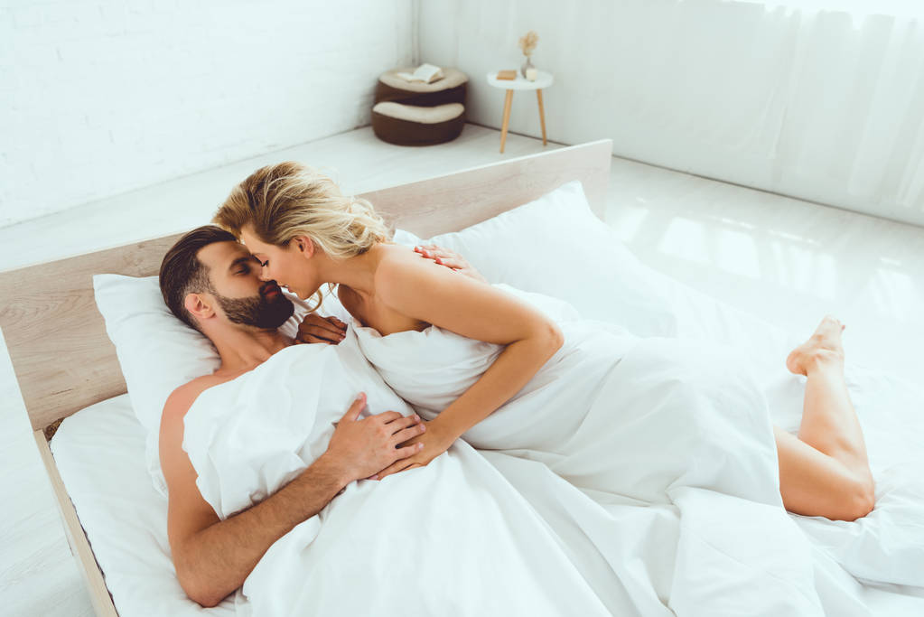 ベッドに横たわっている間、若い女性が彼氏を抱きしめてキスする  - 写真・画像