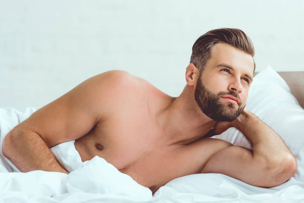 красивый, без рубашки сексуальный мужчина лежит в постели и смотрит в сторону
 - Фото, изображение