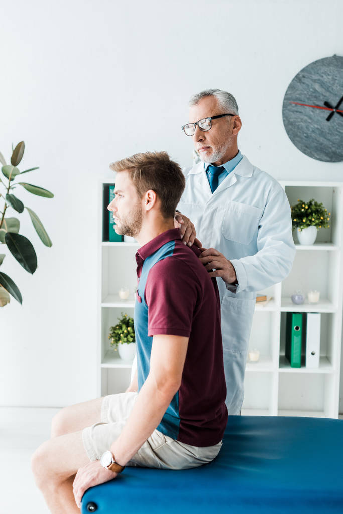 Chiropraktiker in weißem Mantel und Brille berührt Rücken eines gutaussehenden Patienten in Klinik  - Foto, Bild