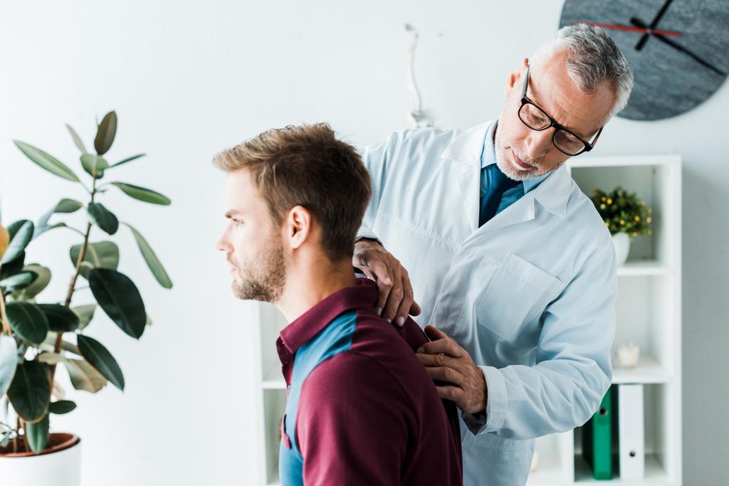 quiropráctico barbudo en bata blanca y gafas tocando la espalda de un paciente guapo en la clínica
  - Foto, Imagen