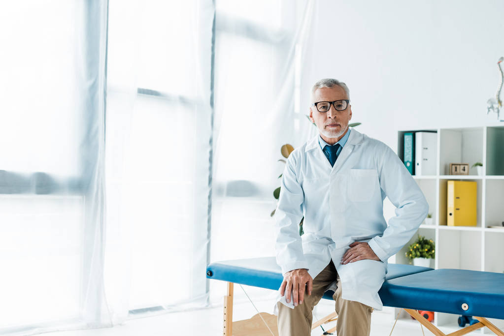 médico barbudo em óculos e casaco branco sentado na mesa de massagem
  - Foto, Imagem