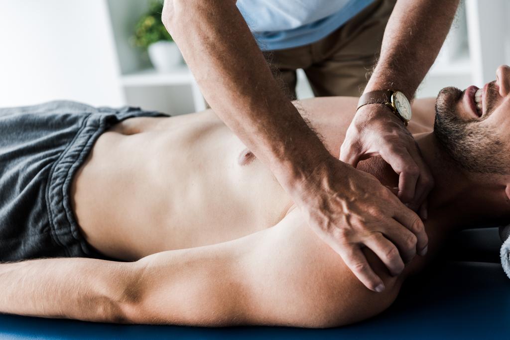 bijgesneden weergave van chiropractor doen massage naar shirtless man met gesloten ogen lijden pijn  - Foto, afbeelding
