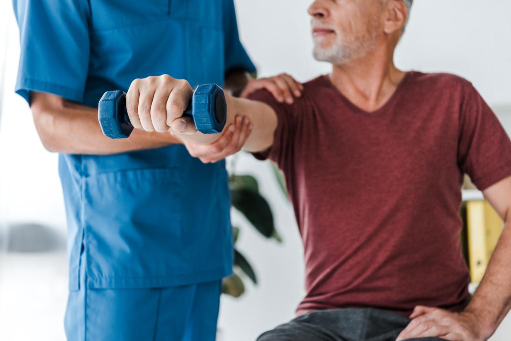 обрезанный вид врача, стоящего рядом с пациентом среднего возраста, упражняющимся с гантелями
  - Фото, изображение