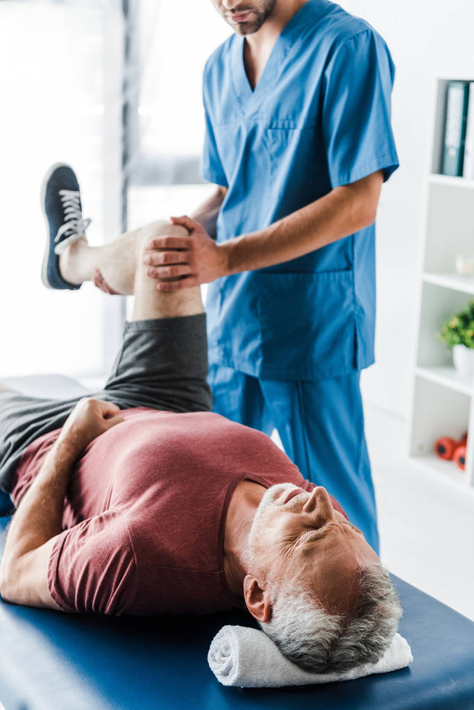 cropped view of doctor touching leg of mature man working out on massage table  - Photo, Image