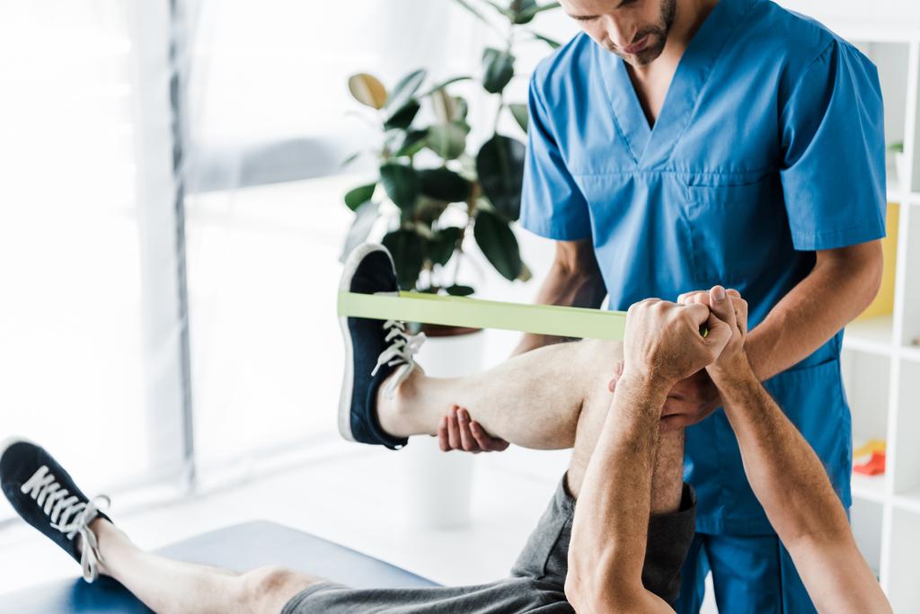 Ausgeschnittene Ansicht eines Arztes, der ein Bein auf einen reifen Mann hält, der mit Spanngurten trainiert  - Foto, Bild
