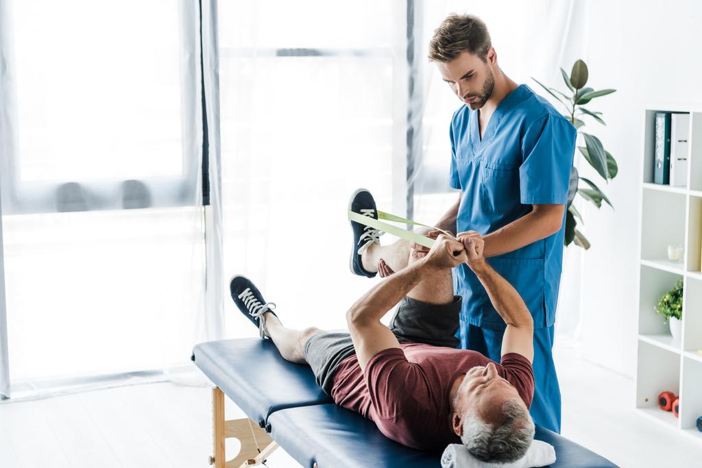 handsome bearded doctor holding leg of mature patient exercising with elastics  - Photo, Image