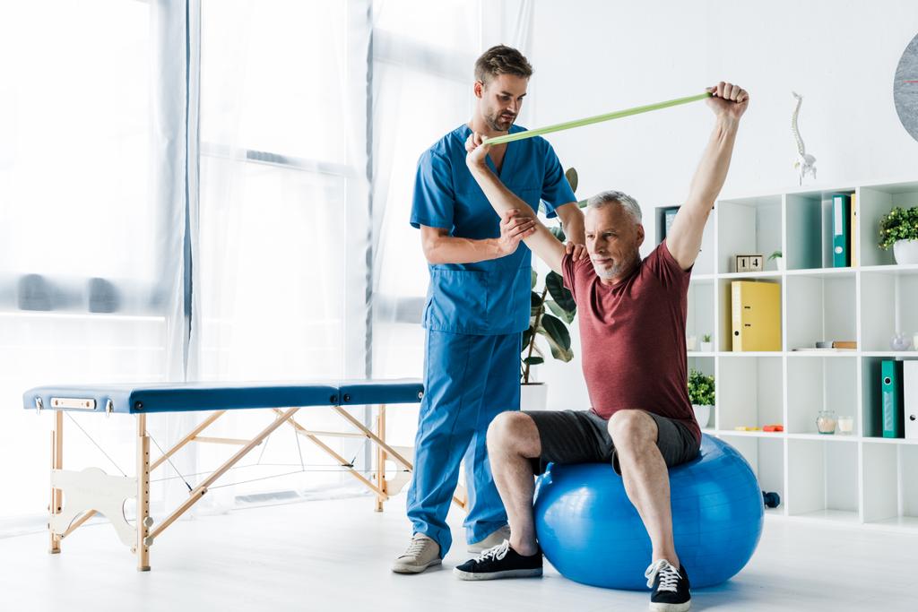 Arzt steht neben Mann mittleren Alters, der auf Fitnessball mit Widerstandsband trainiert  - Foto, Bild