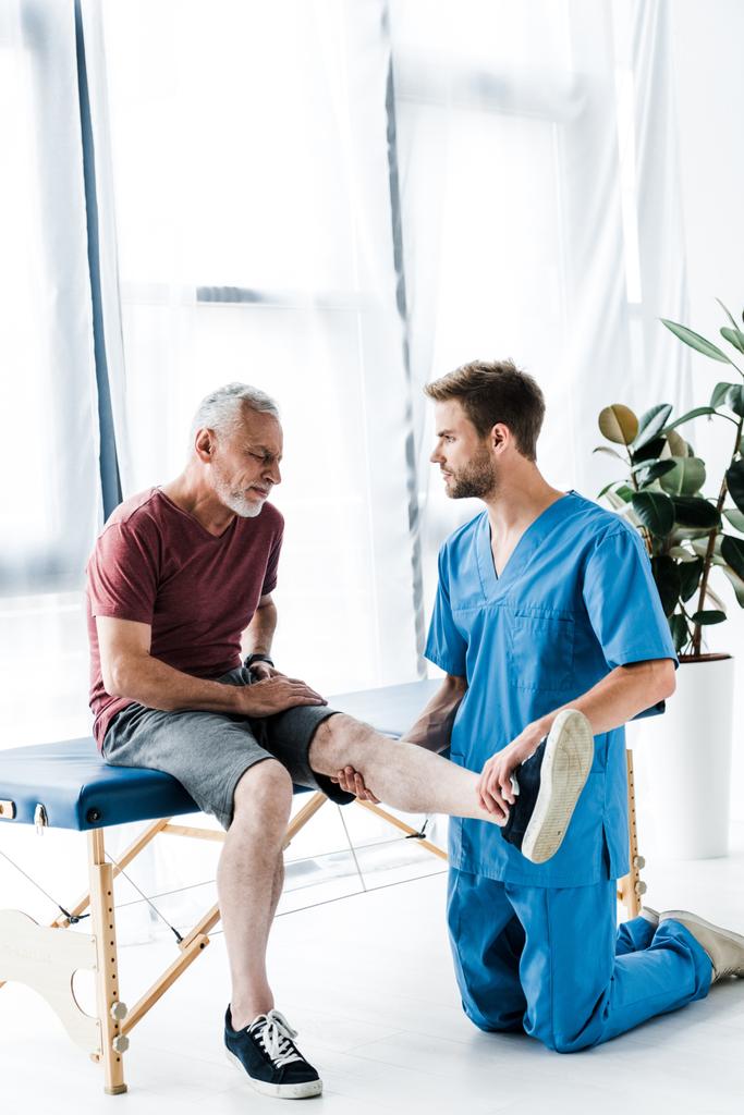 guapo médico tocando pierna y mirando barbudo maduro paciente
  - Foto, Imagen