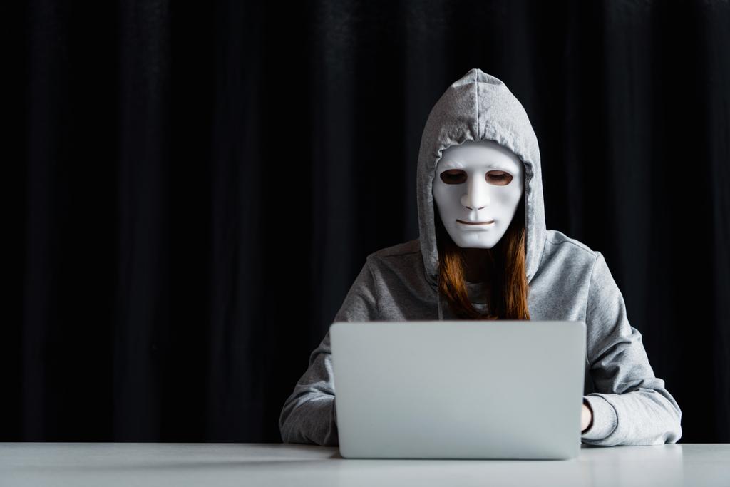 Ανώνυμος Διαδίκτυο τρολ σε μάσκα δακτυλογράφησης στο πληκτρολόγιο του φορητού υπολογιστή στο μαύρο - Φωτογραφία, εικόνα