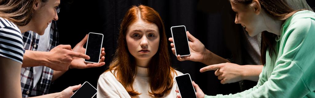 panoráma lövés mosolygó tinédzserek mutatott ujjak a lány közben megfélemlítés és gazdaság okostelefonok üres képernyő fekete - Fotó, kép