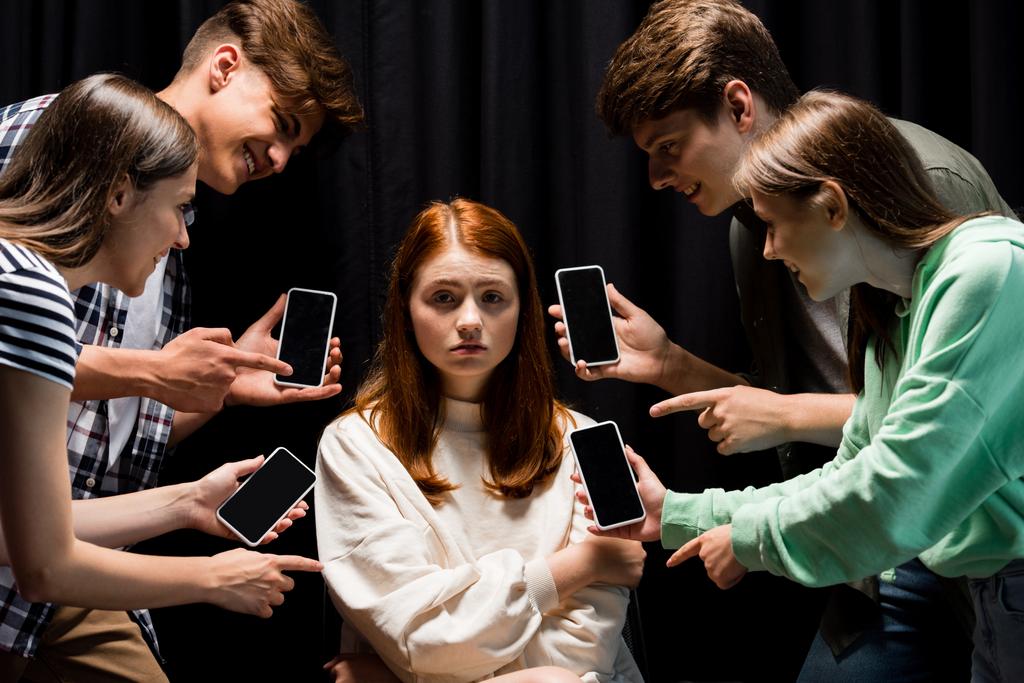 улыбающиеся подростки, указывающие пальцами на девушку во время издевательств и держащие смартфоны с пустым экраном на черном
 - Фото, изображение