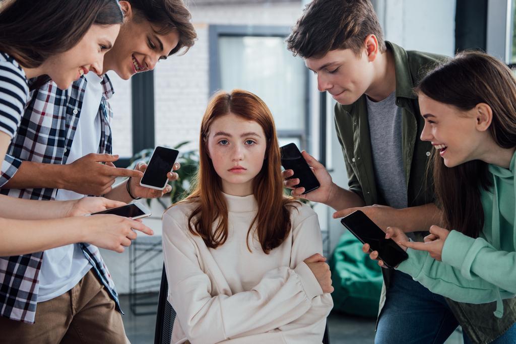 Lächelnde Teenager zeigen beim Mobbing mit den Fingern auf Mädchen und halten Smartphones mit leerem Bildschirm - Foto, Bild