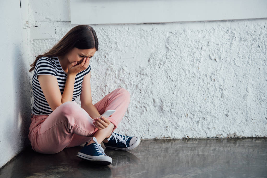 szomorú sír lány rózsaszín nadrágot ül a fal közelében és a gazdaság okostelefon - Fotó, kép