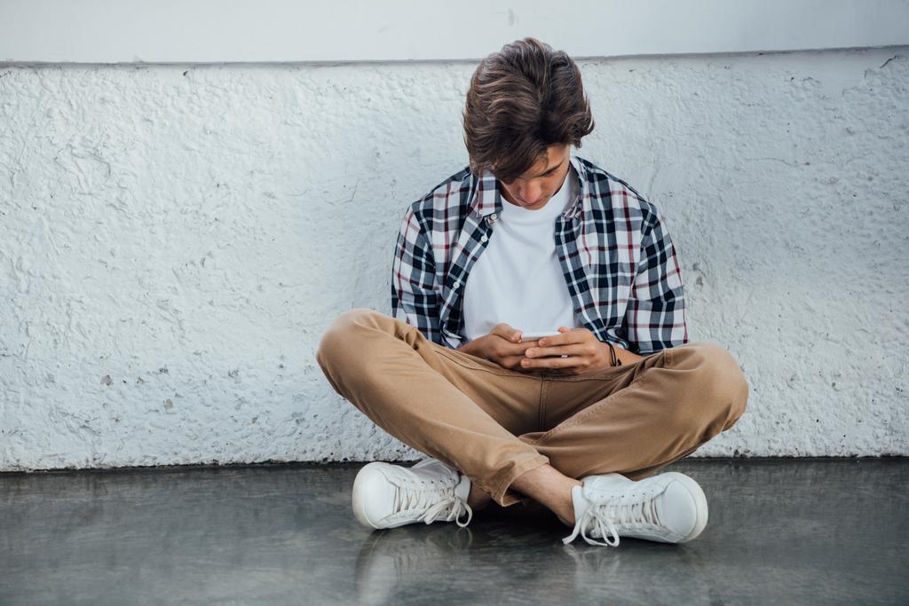 vista frontal del adolescente sentado con las piernas cruzadas y el uso de teléfono inteligente
 - Foto, imagen